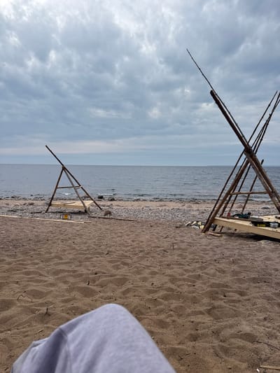 The image depicts a beach scene in Baltiiskoe, Russia. The view is angled, showing the beach in the foreground and the sea and sky in the background. Two wooden structures are visible on the beach. The structures appear to be A-frame or tripod-like, constructed of wooden poles. One structure is taller than the other. The beach consists of sand and small rocks. The sky is overcast with clouds. The sea is calm. Part of a white or gray object, possibly clothing, is visible in the lower right corner of the image. There are no people visible in the main scene.