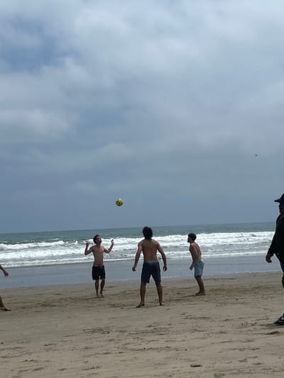 Four men are playing soccer on a beach in Montanita, Ecuador (latitude: -1.825, longitude: -80.755). One man is in mid-air attempting to head the ball. Another man stands on the beach and watches. A man with a black shirt and a black hat is walking away from the scene towards the ocean. The sky is mostly overcast, but there is a small amount of blue showing through the clouds. The sand is brown and wet. There are waves in the ocean behind the men.