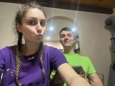 A man and a woman are seen from an upside-down angle, possibly reflected in a mirror on the ceiling or another reflective surface. The man is wearing a bright green shirt and appears to be holding the device taking the photo. The woman is wearing a purple long-sleeved shirt and has her hair in two braids. She is wearing a necklace with a name on it. The background includes a wall, wooden flooring, a pipe, and a hint of an interior space with lights. There is no information to confirm that the image was taken in Vinarovo, Bulgaria.