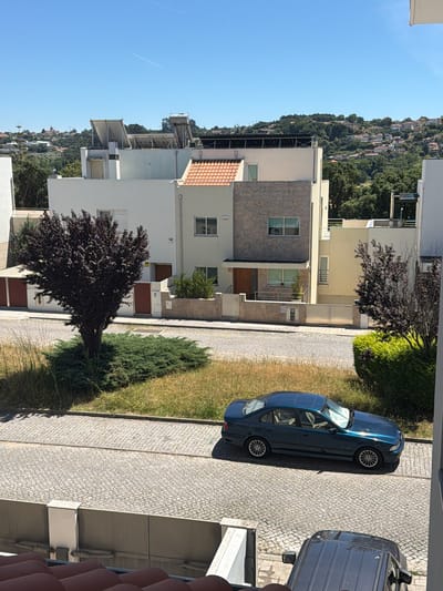 In the city of Gondomar, Portugal, on a clear, sunny day, a two-story modern house is visible from a high vantage point, looking downwards. The house features white and light yellow facades, a stone-clad section, and a red-tiled roof on a small portion of the structure. Solar panels are installed on the flat roof of an adjacent building. In front of the house, a dark blue sedan is parked on a cobblestone street. A dark-colored SUV is partially visible in the foreground, also parked on the cobblestone. Lush green trees and bushes surround the property, and in the background, a hilly landscape is dotted with more houses and trees. The overall scene depicts a residential area in a suburban or semi-rural setting within Gondomar.