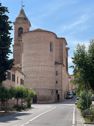In the city of Barbara, Italy, on a sunny day with a clear blue sky, a brick church with a bell tower dominates the scene. The church's rounded apse faces a curving asphalt road with white lane markings. On the left, a tree casts a shadow on the road, partially obscuring a building with closed shutters. A few parked cars are visible further down the road, and on the right, lush green trees and bushes line the sidewalk. The scene captures a quiet moment in the Italian town, with the historic architecture and natural elements creating a picturesque atmosphere.