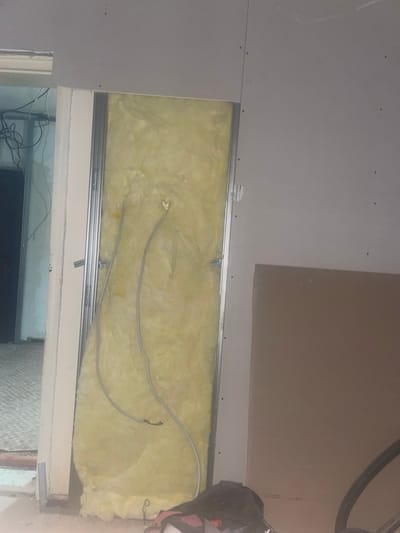 An interior shot depicts a room undergoing renovation. The primary focus is a section of a wall that has been opened up, revealing insulation material, likely fiberglass, in a light yellow hue. This opening is framed by metal studs, commonly used in drywall construction. Electrical wires, thin and gray, are visible within the insulated cavity, suggesting ongoing electrical work.
The surrounding walls appear to be constructed of drywall panels, some of which are already installed and fastened with visible screws, indicated by small dark dots. To the right of the insulated section, a large, flat panel, possibly another piece of drywall or a similar construction board, leans against the wall.
To the left, the scene opens into another area, suggesting a doorway or a larger opening. This area shows more exposed electrical wiring, coiled and hanging loosely. The floor in this adjacent space is partially visible, featuring a patterned tiled surface.
In the foreground, at the bottom of the image, a black bag or backpack rests on the floor, partially obscuring the lower part of the wall opening. The lighting is dim, contributing to the overall impression of an unfinished construction site. The scene is consistent with a residential or commercial building undergoing interior structural and finishing work.
The location provided, Krushevo, Bulgaria, is a geographical reference. While the image itself does not contain explicit visual cues to confirm this specific location, the context of construction materials and practices aligns with typical renovation work that could occur in any urban or semi-urban setting.
