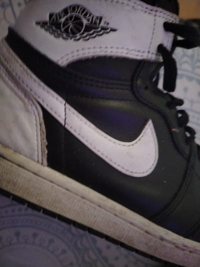 The media file is a close-up, slightly angled shot of a black and white Air Jordan 1 sneaker. The sneaker is the primary subject, with a focus on its side profile and the iconic Nike swoosh. The shoe appears to be made of leather and shows some signs of wear, particularly on the white sole and the white leather accents. The background is a blurry, patterned fabric, possibly a blanket or bedspread. The lighting is somewhat dim, suggesting an indoor setting, and there are no clear indicators of time of day or weather. The sneaker features the Air Jordan logo on the ankle collar, which is a circular emblem with wings and the words "AIR JORDAN" above a basketball. There is also a Nike swoosh logo prominently displayed on the side of the shoe. The image does not depict any people, activity, or specific event. The focus is solely on the details of the sneaker itself. There is no visible text other than the branding on the shoe. The context provided states the location is Bamberg, Germany, but there are no visual cues in the image to confirm this.