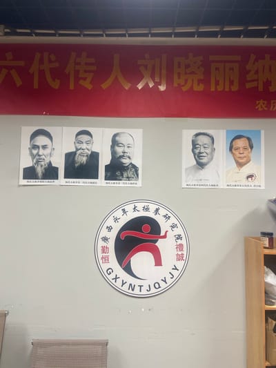 A wall is adorned with a large red banner displaying golden Chinese characters, along with five framed portraits and a circular emblem. The banner's text appears to read "六代传人刘晓丽纳" (Sixth generation inheritor Liu Xiaolijia) and partially visible text on the right side reads "农庆" (Agricultural celebration).
Below the banner, three black and white portraits are arranged horizontally. The first, on the left, shows an elderly man with a long beard and a traditional hat, captioned "杨氏太极拳创始人杨露禅" (Yang Family Tai Chi founder Yang Luchan). The second portrait depicts another elderly man with a long beard and a cap, captioned "杨氏太极拳第二代传人杨健侯" (Yang Family Tai Chi second generation inheritor Yang Jianhou). The third portrait is of a man with a mustache and traditional clothing, captioned "杨氏太极拳第三代传人杨兆瀴" (Yang Family Tai Chi third generation inheritor Yang Zhaoying).
To the right of these, two color portraits are also arranged horizontally. The first shows an elderly man in a white collared shirt, captioned "杨氏太极拳第四代传人杨振基" (Yang Family Tai Chi fourth generation inheritor Yang Zhenji). The second portrait, to its right, shows a man wearing a white martial arts uniform, captioned "杨氏太极拳第五代传人洪日健" (Yang Family Tai Chi fifth generation inheritor Hong Rijian).
Centered below the portraits is a large, circular emblem with a red and black yin-yang-like symbol in the center, depicting a stylized human figure in motion. The emblem is surrounded by Chinese characters and English lettering that appears to read "GXYNTJ QY".
The wall is painted a light gray. To the right, a wooden shelving unit is visible, with various items including a jar with a red label on the top shelf and cardboard boxes on the bottom. Two chairs with striped cushions are positioned in the lower left corner of the image. The overall setting suggests an indoor space, possibly a martial arts school or a cultural exhibition hall.
Based on the context of the characters and the style of the display, this image is likely located in Nanning, China, potentially within a facility dedicated to the study or promotion of Yang Family Tai Chi.