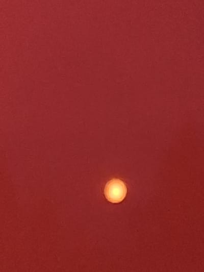 The image is a close-up, abstract shot of a red surface with a single, small, glowing yellow-orange orb positioned in the lower center. The orb appears to be a light source, possibly a lamp or a star, with a soft halo effect. The red surface has a textured appearance, suggesting it might be fabric, painted wall, or paper. There are no discernible features of the city of Lesouchastok 831, Russia in this image; it is solely focused on the abstract composition of the red background and the central light source.