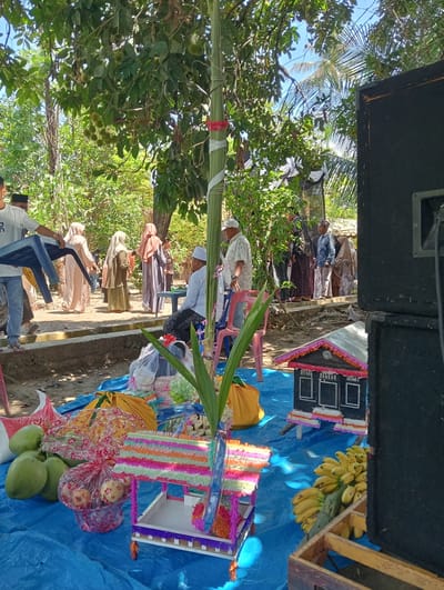 A bright, sunny outdoor scene in Peudari, Indonesia, depicts what appears to be a cultural gathering or ceremony. A group of people, many wearing traditional Islamic clothing such as headscarves and long robes, are visible in the background, standing or walking amongst trees. In the foreground, a blue tarp is spread out on the ground, laden with offerings and decorative items. These include several green coconuts, a basket of red apples, and bunches of bananas. Two small, colorful, miniature structures, resembling houses or stalls, are placed on the tarp, adorned with intricate, layered decorations. A large, black speaker stands to the right of the frame, suggesting a musical or public address component to the event. A tall, bamboo-like pole, wrapped with white and red ribbon, rises vertically on the left side of the image, a prominent ceremonial element. The overall impression is one of a communal celebration taking place in a tropical, natural environment.