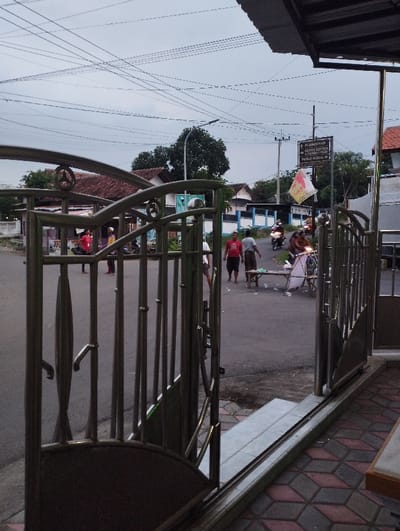 The image captures an everyday street scene in Kecamatan Panji, Indonesia, viewed through an open, ornate metal gate. The gate, likely made of stainless steel, features vertical bars and decorative curved elements. In the immediate foreground, a tiled pavement with a geometric pattern is visible to the right.
The midground reveals a paved road with several people and motorcycles. Some individuals are standing on the street, while others are riding motorcycles, indicating typical daily activity. To the right of the street, a small table or bench is present, near what appears to be a partially visible street vendor's cart or a structure with a white tarp.
In the background, traditional houses with reddish-brown tiled roofs are scattered amongst lush green trees under an overcast sky. Numerous overhead power lines crisscross the sky, adding to the urban landscape. A street light pole is also visible.
A prominent brown sign on a pole stands on the right side of the road. Visible text on the sign includes directional information: "HLARIPANAN", "DUSUN COTTON", "RUMAH PAK LURAH" (Head of the Village's House), and "PANJI KIDUL" (South Panji), all with arrows pointing to the right. A small, colorful flag is attached to the sign pole.
The overall lighting suggests either early morning, late afternoon, or a heavily cloudy day, as there are no harsh shadows. The atmosphere appears calm, depicting a routine moment in a residential or semi-rural area.