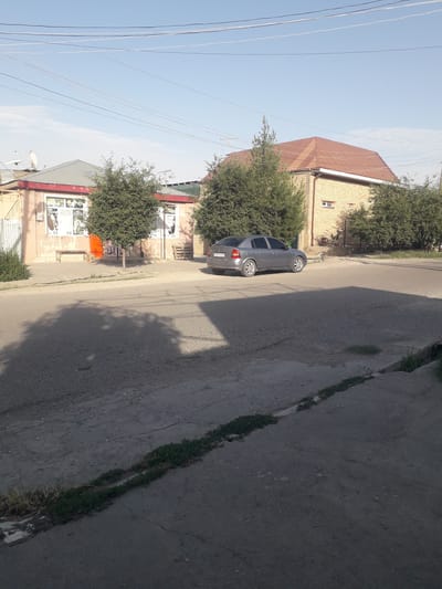 The image displays a sunny, daytime street scene in Khistevarz, Tajikistan. A dark grey sedan, possibly a Chevrolet Aveo or a similar model, is parked facing away from the viewer on the left side of a paved road. The car features distinctive silver multi-spoke wheels, and a portion of its license plate, "01," is visible on the rear.
Along the left side of the street, several buildings are present. One notable structure has light-colored brick walls and a reddish-brown tiled roof, with a white-framed window. Further down the street, another building with a red roofline features lighter-colored walls and large windows, possibly storefronts. One window display contains faint, largely unreadable text, with a fragment resembling "EARTH" being vaguely discernible amidst other graphics.
Numerous green, leafy trees and bushes line the road and are interspersed around the buildings, casting shadows onto the street. Above, multiple white power lines stretch across a clear, light blue sky, which shows some faint, wispy clouds. The asphalt road appears worn with cracks, particularly a visible linear crack along the right side where some grass and weeds have grown. No people are visible in the scene, which conveys a calm and quiet atmosphere, typical of a residential or commercial street. The shadows suggest the time of day is likely late morning or early afternoon.