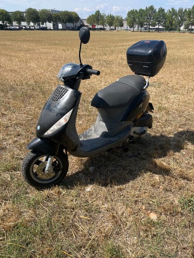 A black Piaggio Zip scooter is parked on dry, brown grass in an open field. The scooter has a black top case mounted on the rear. In the background, a row of green trees stands in front of a line of buildings, likely industrial or commercial. Cars are parked in front of the buildings. The sky is partly cloudy and blue. The location is Castiglione delle Stiviere, Italy.