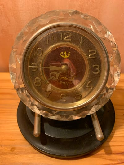 An antique alarm clock is displayed on a wooden surface, with a textured orange wall in the background. The clock has a round, brass-colored face with Roman numerals and decorative hands. The glass cover of the clock has a faceted, crystal-like border. The clock is supported by two silver-toned legs that rest on a black, circular base. A logo featuring a stylized crown and Cyrillic lettering is visible on the clock face, as well as text that appears to read "Jemala". The overall condition of the clock suggests it is an older item. The scene is lit with warm, artificial light. The specific location in Mepiskalaki, Georgia, is not determinable from the image alone.