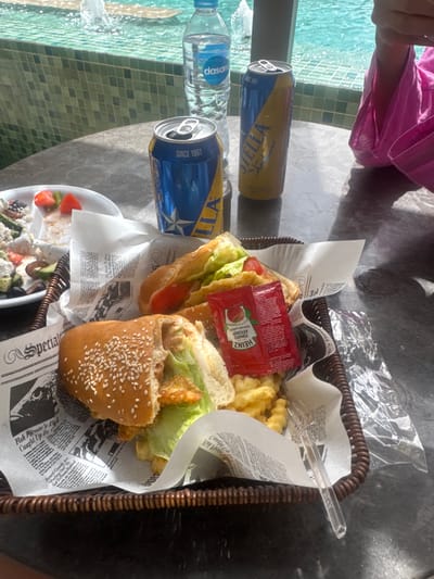 Captured from an aerial perspective, the image presents a scene from El Gouna, Egypt, beside a pool. A person, partially visible in a pink top, is present. The focal point is a brown woven basket containing a partially consumed burger with sesame seeds on the bun, accompanied by lettuce, tomato, and what appears to be fried chicken. The basket also holds French fries and two packets of Heinz tomato ketchup. The food is served on newspaper-print lining. A small plate with salad containing feta cheese and black olives sits near the basket. A bottle of Dasani water and two cans of Stella beer are placed beside the basket. The pool features green tiles.