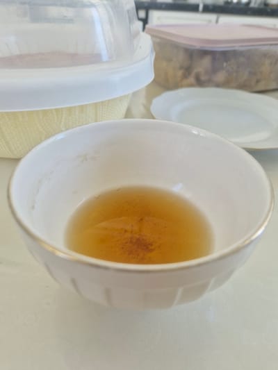 A close-up, top-down shot shows a white ceramic bowl with a gold rim, containing a small amount of amber-colored liquid. Some reddish-brown sediment is visible at the bottom of the bowl. In the background, out of focus, are kitchen items including a clear plastic container with a pink lid filled with food, and a white plate. The scene appears to be indoors, likely a kitchen counter. There is no indication of the city of Zhanaozen, Kazakhstan, or any people in the image.