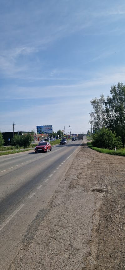 The image depicts a roadside scene in Leninskoye, Russia, on a clear day. A two-lane asphalt road runs through the frame, with a red car driving in the left lane and other vehicles, including a truck and several cars, visible further down the road. The road is bordered by a gravel shoulder on the right and vegetation, including trees and bushes, on both sides. A billboard advertising "Sun Supermarket" is visible on the left, along with some buildings and utility poles. The sky is blue with scattered white clouds. The overall impression is a typical rural or suburban road with moderate traffic.