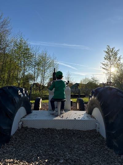 The image, when viewed in correct landscape orientation, captures an outdoor scene under a bright, clear blue sky. A child, seen from behind, is the primary subject, wearing a green hoodie, a green cap, dark trousers, and white sneakers. They are positioned on a metallic, diamond-plate textured platform, engaged in operating a black, cannon-like play device.
This platform appears to be part of a larger, robust structure, possibly a playground apparatus designed to resemble a vehicle or tractor, featuring prominent, dark, multi-ridged tires or fenders. The ground immediately surrounding this structure is covered in dark wood chips or mulch.
In the background, numerous trees with vibrant, light green leaves are visible, suggesting a spring or early summer season. The sun is a radiant point in the upper right quadrant of the sky, casting bright rays and some lens flare, indicative of either morning or late afternoon. Several faint contrails trace paths across the vast blue sky. The overall setting appears to be an outdoor recreational area or playground, likely in Olingen, Luxembourg, where the child is immersed in play. Faint, indistinct text is visible on the side of one of the large dark structures.