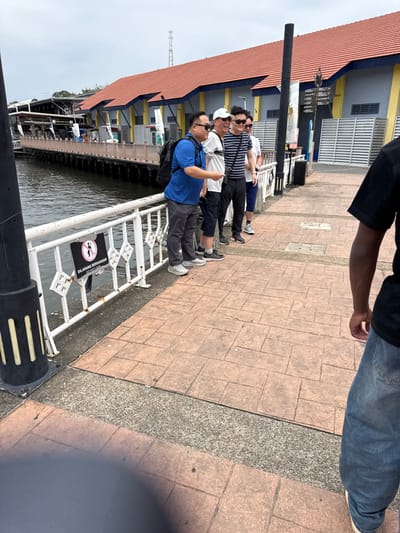 Four men stand on a dock next to a body of water. The dock has a white railing with a sign in a black circle that says "Dilarang Passangar". The man closest to the camera wears a blue shirt and black backpack. The next man wears a white shirt, and the two men behind him wear striped shirts. The ground is covered in brick. A man in jeans and a black shirt stands facing the camera.