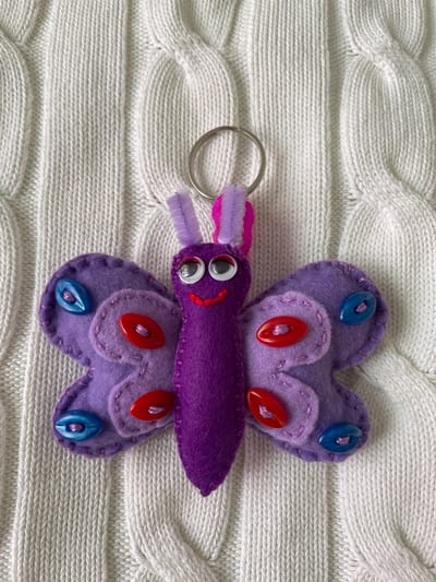 A top-down shot shows a handmade purple butterfly keychain on a white knitted background. The butterfly's wings are heart-shaped and adorned with blue and red buttons. The body is a darker shade of purple, with googly eyes and a small red smile. A purple and pink fuzzy pipe cleaner forms the antennae. A silver keyring is attached to the butterfly. The knitted background has a cable-knit pattern.
