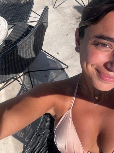 The scene depicts a female individual outdoors in Mykonos, Greece, under direct sunlight. The subject is captured in a close-up, high-angle selfie, showing her right arm, upper torso, and the right side of her smiling face. She has tanned skin, dark hair pulled back, and is wearing a light peach-colored bikini top. Her visible jewelry includes a single silver stud earring in her right earlobe and two delicate gold-colored necklaces: a lower chain with a small, round, clear pendant, and a higher chain with a small gold bear-shaped pendant.
To the left of the subject, two dark, possibly black, wicker or rattan chairs with slim black metal frames are visible. A small, light-colored, circular tabletop, possibly stone, is partially present at the far left. The ground is composed of light-colored square tiles or paving stones. Strong shadows are cast from the chairs and the subject, indicating bright sunlight from a high angle. Scattered small green leaves are visible on the tiled surface.