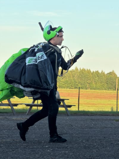 A person wearing a black skydiver's suit, a bright green helmet with a clear visor, and black gloves is walking from left to right across a gravel surface. They are carrying a large, dark-colored parachute bag with green fabric visible underneath. The person is looking to the right, with their right arm extended. In the background, a wooden picnic table is visible, beyond which is a field of grass and trees. A fence line with orange netting runs through the field. The sky is clear and pale blue. The scene appears to be an outdoor airfield or drop zone. The location is identified as Limbaži, Latvia.