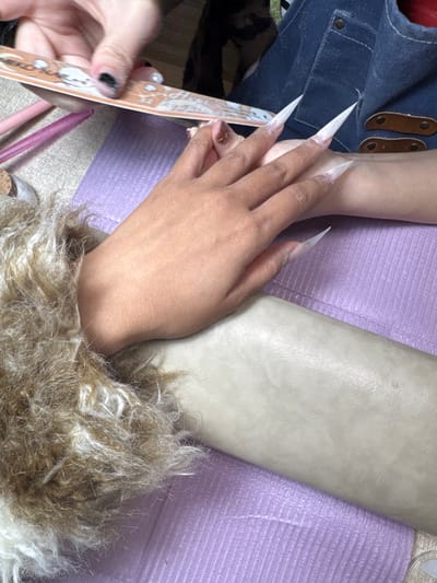 A person files another's long, pointed nails with a file. The person receiving the manicure has a tan hand and forearm. They are wearing a fuzzy sweater. The manicurist has black nail polish. A denim shirt is visible, as well as an off-white leather surface and a lavender textured surface. Pink manicure sticks are also visible.