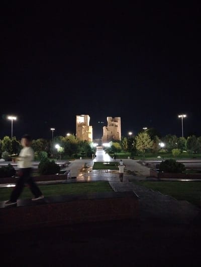 The image depicts a night scene in Shahrisabz City, Uzbekistan, featuring the Ak-Saray Palace ruins. The ruins, illuminated by artificial lighting, are the central focus in the mid-ground. In the foreground, to the left, a person is captured in motion, appearing as a blurred figure walking from left to right. To the right, a second person is visible, also in motion, facing away from the camera and walking towards the ruins. The environment includes manicured lawns, paved walkways that appear wet and reflective, and trees. Streetlights are visible in the background, illuminating the area and contributing to the overall nighttime ambiance. The sky is dark, indicating it is night.