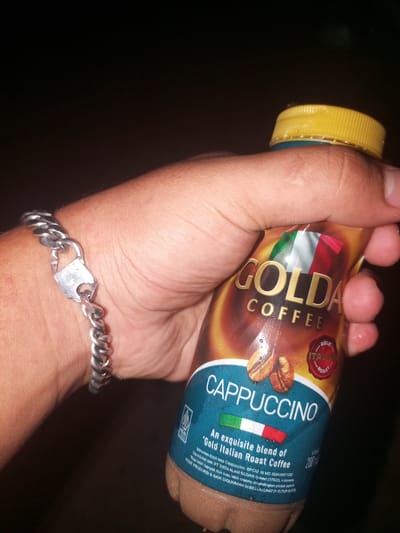 A hand is holding a bottle of Golda Coffee Cappuccino, highlighting its Italian roast blend. The bottle has a yellow cap, and the arm has a silver chain-link bracelet with a square clasp. The background is dark. The label states the coffee is produced by PT Tirta Alam Segar in Bekasi (17520), Indonesia. Lawang, Indonesia is not directly mentioned in the context of the image.