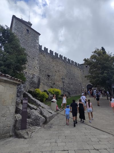 The image depicts a stone fortress wall under a cloudy sky, likely in the City of San Marino. A group of people, including families with children, are walking up a paved pathway leading towards an arched entrance in the wall. The path is lined with trees and some shrubbery.

The subjects are diverse, with individuals of various ages and genders dressed in casual summer clothing. Some people are walking ahead, while others are walking together in small groups. Their postures and the direction of their movement suggest they are tourists exploring the historical site. The atmosphere appears relaxed, with a sense of exploration and leisure.

Notable details include the weathered texture of the stone fortress, the crenellated battlements along the top of the wall, and a single window visible on a tower. A lamppost stands beside the pathway, adding to the historical ambiance. A dark sign with some text is placed on a rocky outcrop to the left of the path. The lighting suggests it is daytime, possibly late afternoon, given the soft light and overcast conditions.

The location is identifiable as the City of San Marino due to the distinctive architecture of the fortress and the surrounding landscape. The presence of tourists strongly indicates this is a popular historical attraction. The overall scene conveys a sense of historical grandeur combined with contemporary tourism.