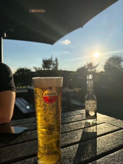The image displays an outdoor scene at a drinking establishment in Brandesburton, United Kingdom, captured during late afternoon or early evening under direct sunlight. In the immediate foreground, a tall glass of Amstel Bier, filled with golden liquid and topped with a white foam head, stands prominently on a dark, slatted outdoor table. Small bubbles are visible within the beer. To the right of the glass, an empty, clear glass bottle of Corona Extra beer rests upright on the same table, its label facing forward. Direct sunlight from the right side of the frame creates strong highlights and long shadows across the table surface. A partial view of a person, consisting of a left arm and shoulder clad in a dark short-sleeved shirt, is visible on the far left, out of focus. The upper left portion of the frame is shaded by a large, dark canopy or umbrella. The background features blurred wooden outdoor tables or benches, distant green foliage (trees and bushes), and a bright blue sky with a few wispy white clouds. The sun is visible as an intense circular light source low in the sky on the right side, illuminating the scene.