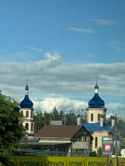 The image depicts an outdoor daytime scene on Peremohy Street, Ukraine, under a bright blue sky partially obscured by white and grey cumulus clouds. The primary subject is an Orthodox church complex featuring multiple domed structures. Two prominent cupolas are visible, both topped with reflective blue domes and gold-colored Orthodox crosses. The main body of the church is constructed from light-colored brick or stucco, accented with reddish-orange trim around its arched windows and cornices. Several smaller blue domes with crosses are also present towards the right and rear of the main structure.

In front of the church, slightly to the left, stands a building with a brown, steeply pitched roof and dark wooden siding, obscuring the lower section of the church. Additional wooden structures are visible adjacent to it. A line of coniferous trees is present in the background behind the buildings.

The lower foreground is dominated by a large yellow banner or sign displaying Ukrainian text, partially legible as "ДОБРИВА ГРУНТИ ПОЛИВ РОСЛИНИ" (Fertilizers, Soil, Irrigation, Plants), indicating an agricultural or garden supply business. To the far right, a vertical pole supports a rectangular sign featuring a green "ФОРА" (Fora) logo. White text on this sign indicates operating hours "з 6:00 до 23:00" (from 6:00 to 23:00) and an address: "м. Київ, вул. Кудряшова, 44Б" (Kyiv, Kudryashova Street, 44B). No individuals are visible within the frame. The scene is well-lit by natural daylight.