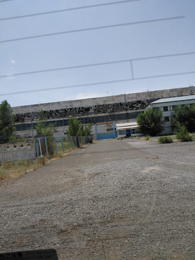 The image shows a long, weathered industrial building with many broken windows, suggesting it is abandoned or derelict. In front of the building, there is a gravel road leading towards a blue gate and a smaller building. A chain-link fence with a light blue top runs along the left side of the road, separating it from a low concrete wall. Trees and dry grass are visible on the left side of the frame. The sky is cloudy and overcast. The overall scene appears to be in a state of disrepair, possibly within an industrial area of Ақтүбек, Kazakhstan. There are no people or animals visible in the image.