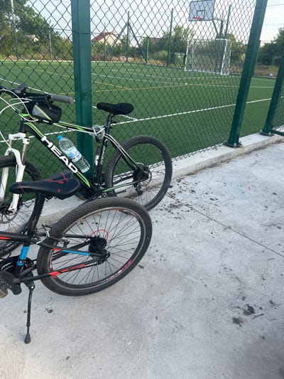 The image depicts two bicycles lying on a concrete surface alongside a green chain-link fence. Beyond the fence is a green artificial turf field with white lines, suggesting a sports field. A basketball hoop and soccer goals are visible in the background. One bicycle is predominantly black and white with green accents, branded with "HEAD" on the frame. A water bottle is attached to the bike frame. The second bicycle is mostly black with blue and red accents. The location appears to be in close proximity to a sports facility, possibly in Kalchevo, Bulgaria. There are no people visible in the image.