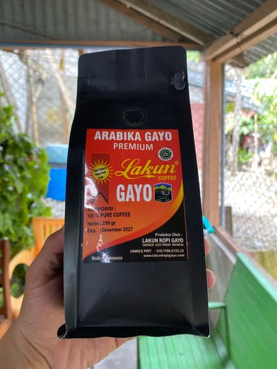 A person's hand is holding a black, upright coffee bag in front of a blurry outdoor background that suggests a residential area or covered outdoor space with some vegetation and a green bench. The coffee bag features prominent text and graphics. At the top, it reads "ARABIKA GAYO PREMIUM." Below this, a sunburst graphic contains the text "Masi Seperti Yang Anda Suka" (Still Like You Like). The brand name "Lakun COFFEE" is displayed in large, stylized lettering. Further down, "GAYO" is written in large, bold letters, accompanied by a smaller image of a landscape with a bird. Information about the product includes "KOMPOSISI: 100 % PURE COFFEE," "Netto: 250 gr," and "Exp.: Desember 2027." At the bottom, it states "Made In Indonesia" and lists production details: "Produksi Oleh : LAKUN KOPI GAYO TAKENGON - ACEH TENGAH - INDONESIA" and "DINKES PIRT : 510.1106.0155-22 www.lakunkopigayo.com." The setting does not provide specific visual cues to pinpoint the exact location within Suakbilie, Indonesia, but the overall impression is a casual, everyday scene.