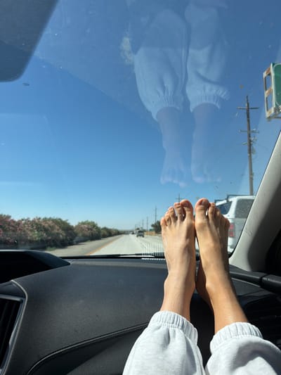 In Bakersfield, California, a person's bare feet are resting on the dashboard of a car, with their reflection visible on the windshield. The car is on a highway, with cars driving in the distance and flowering bushes lining the road. The sky is clear and blue.