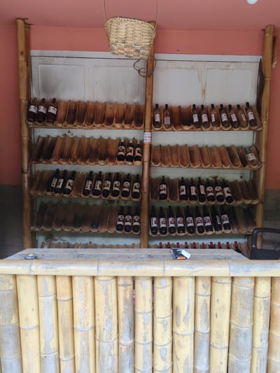 The image depicts a display of bottled beverages, likely wine or spirits, arranged on tiered shelves made of bamboo. The shelves themselves are designed with carved bamboo segments that cradle the bottles. The backdrop appears to be a white, paneled wall, possibly a door or cabinet. Above the shelves, a woven basket hangs suspended. The entire structure is framed by vertical bamboo poles. In the foreground, a bar constructed from vertical bamboo stalks is visible, with a wooden countertop on which a small black device with an orange button rests.

The lighting suggests it is daytime, and the setting appears to be an indoor or covered outdoor area, given the wall and the lack of natural light indicative of open sky. The overall aesthetic is rustic and natural, utilizing bamboo extensively in both the display and the bar. The bottles are varied in color and labeling, some appearing dark, others lighter, suggesting different types of beverages. There is no discernible activity or people in the image.

Some faint text is visible on labels on the bottles, but it is too indistinct to read. A small label with "VISA" is visible on the left side of the middle shelf. The location context of Pirveli Maisi, Georgia, suggests this might be a local winery, distillery, or a bar with a focus on regional products.