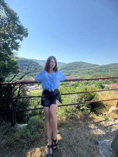 A woman is posing for a photo while leaning against a metal railing. She is wearing a light blue short-sleeved shirt tied at the waist and a black leather skirt. She has dark hair and appears to be looking at the camera. Her legs are crossed, and she is wearing black sandals. The background shows a lush green mountainside covered in trees, with a clear blue sky above. There are some buildings visible in the distance on the mountainside. The scene appears to be outdoors during the daytime, with bright sunlight creating shadows. The woman's pose and expression suggest she is relaxed and enjoying the view. The presence of the railing and the elevated perspective suggest she is on a viewpoint or a scenic overlook. The vegetation and the style of the buildings hint at a European setting.