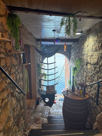 The image captures a narrow passageway in the city of Grad Rovinj, Croatia. The passage is enclosed by old stone walls. At the entrance is a large wine barrel that serves as a table, and a black metal fish skeleton sculpture is suspended overhead. In the distance, a glimpse of the Adriatic Sea can be seen, framing the view. Vegetation is visible throughout. A person is visible in the background.