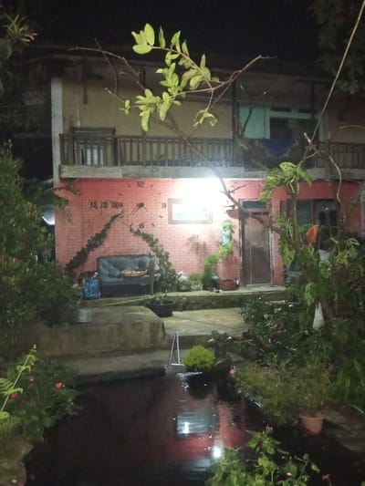 The image displays a nighttime outdoor scene in Naya Andura, India, featuring a two-story building and a water feature. The ground floor of the building is constructed with red brick, partially covered by climbing vines, and prominently features a decorative wall clock with numbers 12, 3, 6, and 9 visible. To the left of a brightly illuminated rectangular window, an outdoor sofa with grey cushions rests, with a blue bag positioned beside it. A wooden door is located to the right of the window, surrounded by multiple potted plants and hanging foliage. The upper floor is lighter in color with a wooden balcony railing.
In the foreground, a dark, reflective body of water, possibly a pond or pool, occupies the lower portion of the frame, reflecting artificial light sources. Stone steps or raised platforms separate the water from the building. Abundant green foliage, including various potted plants, is distributed around the water feature and along the path leading to the building. A small, light-colored, ladder-like metallic structure is partially submerged in the water, adjacent to a small, round potted plant. Overhead branches with light green leaves partially frame the top of the image. A strong artificial light source illuminates the ground floor wall and portions of the surrounding vegetation. No individuals or explicit actions are discernible.