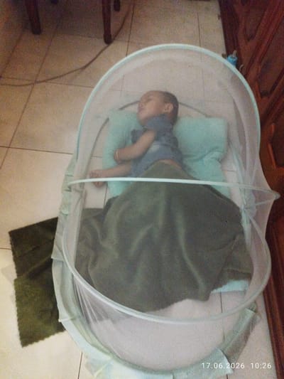 The image shows a young child sleeping in a mosquito net cot on a tiled floor. The child is wearing a blue shirt and is covered with a dark green blanket. There are two light blue pillows under the child's head. A dark green mat is visible to the left of the cot. In the background, there is a wooden cabinet and a glimpse of a cord on the floor. The timestamp at the bottom right of the image indicates June 17, 2026, at 10:26. The scene appears to be an indoor residential setting, likely a bedroom. There is no specific indication of the city of Kampot, Cambodia, within the image itself.