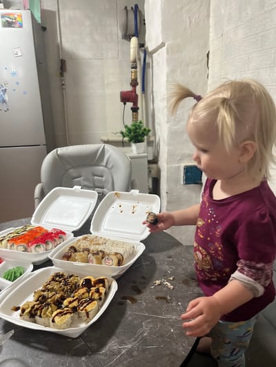 A young Caucasian girl with blonde hair pulled into two pigtails stands in front of a table laden with several containers of sushi. She is wearing a maroon long-sleeved shirt with a graphic print and blue patterned leggings. She holds a single piece of sushi in her right hand, examining it. The table is dark, with a marble-like pattern. Behind the table, a grey high chair is visible. To the left, part of a silver refrigerator is seen, adorned with stickers. The background features a white brick wall and industrial-looking piping. The setting appears to be a domestic environment, possibly a kitchen or dining area, located in Rakhinka, Russia.