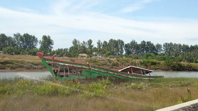 The image depicts a weathered, green and red wooden boat resting on dry land near a body of water, with a lush green tree line forming the background. The boat is oriented diagonally from the lower left to the upper right of the frame. Its hull is visibly aged, with some sections appearing to be missing or damaged. The interior of the boat is exposed, revealing wooden supports and framing. A white canopy structure with red trim is present on the rear portion of the boat. The surrounding environment consists of dry, tall grass and vegetation. In the distance, across the water, a small settlement with several buildings, including some with yellow roofs, is visible nestled amongst more trees. The sky is partially cloudy, with patches of blue. The scene appears to be a waterfront area in or near Tanjong Krueng, Indonesia. There are no people visible in the image.
