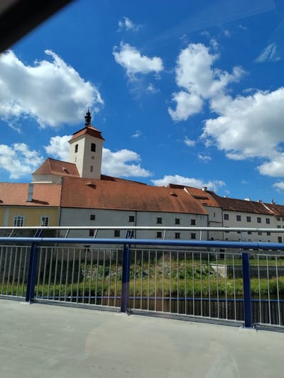 The image shows a view of the Strakonice Castle complex in Strakonice, Czechia. In the foreground, there is a concrete walkway with a blue and silver metal railing, obscuring the lower portion of the castle. Behind the railing, a body of water with a grassy bank is visible. The castle itself features red-tiled roofs and white-painted walls. A prominent bell tower with a dark spire rises above the main structure. The sky is blue with scattered white clouds. The scene appears to be captured on a sunny day.