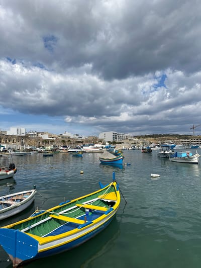 The image depicts a harbor in Marsaxlokk, Malta, under a cloudy sky. Several traditional Maltese fishing boats, known as 'luzzus,' are docked in the water. The luzzus feature vibrant colors, including blue, yellow, and green. Buildings line the waterfront, ranging from low-rise structures to modern white buildings. A construction crane is visible in the background. The water is relatively calm, with slight ripples on the surface. There are some visible objects floating on the water surface. The scene is devoid of people.