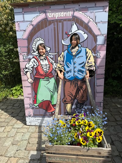 Two children stand behind a photo cutout board featuring painted figures of a man and a woman dressed in traditional clothing. The children have placed their faces through holes in the board to look like they are part of the painted figures. The man is in a blue jacket and hat while the woman is in a red and green dress with a white bonnet. Both figures are holding wooden sticks. The board has "Gripsdrill" written on the side. To the right of the board is a wooden box filled with colorful pansies and forget-me-nots. The scene is located outdoors on a stone-paved surface in Cleebronn, Germany.