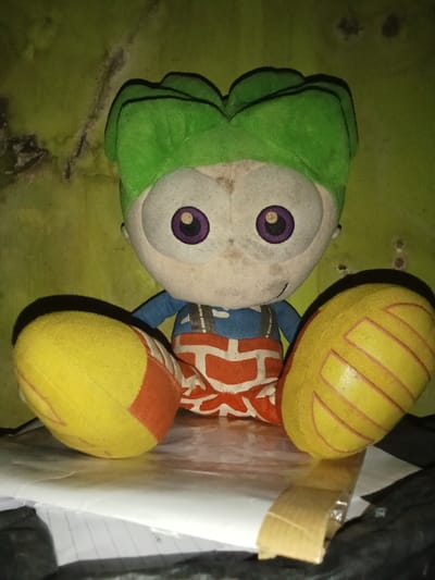 A soiled plush toy is depicted in a close-up, frontal view, situated in an indoor environment in Ciputri, Indonesia. The toy features a beige face with prominent white eyes and purple irises, a green leaf-like headpiece, a blue torso, and orange pants with white geometric patterns. Its two large, yellow, oval-shaped appendages (legs/feet) are splayed outwards, each accented with orange and faint yellow stitching. The toy rests on a stack of white paper, partially obscuring a dark, possibly fabric or plastic, object in the lower right foreground. A piece of brown adhesive tape is visible on the paper to the right of the toy. The background consists of a light green wall exhibiting significant dark discoloration and staining, consistent with dampness or mold. No people are visible.