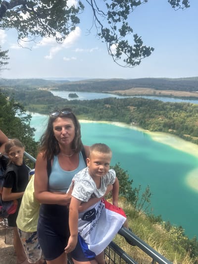 This media file captures a family enjoying a scenic overlook on a sunny day. The subjects are a woman and three children, two boys and a girl. The woman, positioned in the foreground, is wearing sunglasses on her head and a light blue tank top. She is holding one of the young boys, who is wearing a grey and white striped shirt. To her right, another child, a girl, is partially visible wearing a black shirt. Further down, another young boy is visible.
The group is standing near a metal railing, suggesting they are on a viewing platform or at a scenic lookout. Behind them, a breathtaking landscape unfolds. A large body of turquoise water, possibly a lake or reservoir, dominates the background, with lush green forests and hills surrounding it. The sky is clear and bright, indicating daytime with good weather. The image is oriented vertically, with the landscape appearing to stretch out above the people.
There is visible text on the white and blue flag held by one of the children, but it is partially obscured and difficult to read clearly. The overall mood appears to be joyful and relaxed, as the family takes in the beautiful view. The location context of La Chaux-du-Dombief, France, suggests this could be a popular tourist spot known for its natural beauty.