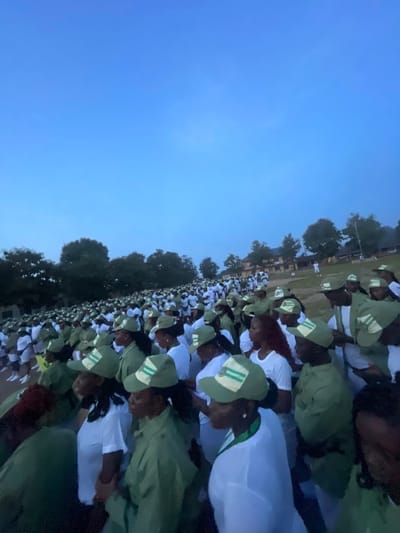 In the city of Ise - Ekiti, Nigeria, a large gathering of people, predominantly young adults, are assembled outdoors. They are dressed in matching light green uniforms with white accents and caps, suggesting an organized event or program. The individuals are standing close together, facing forward, and appear to be engaged in an activity, possibly a ceremony or a briefing. The background features a line of trees and buildings, indicating a semi-rural or institutional setting. The sky is a clear blue, suggesting it is daytime. The composition of the image shows a wide shot, emphasizing the scale of the group.