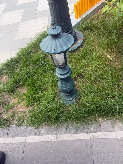 This image depicts a streetlamp situated in a grassy area adjacent to a paved walkway. The streetlamp is dark green with ornate detailing and a glass enclosure. It stands near a larger dark green pole, possibly another lamppost or part of a structure. The grass is green and somewhat uneven, with patches of drier soil visible. The walkway is made of grey stone pavers laid in a geometric pattern, with a lighter colored border. In the background, a row of yellow posts is visible behind a low grey wall. The scene is likely located in a public area in the city of Aihui, China, given the context provided. There are no people clearly visible in the frame, nor any specific actions taking place. The overall impression is one of an urban outdoor setting.