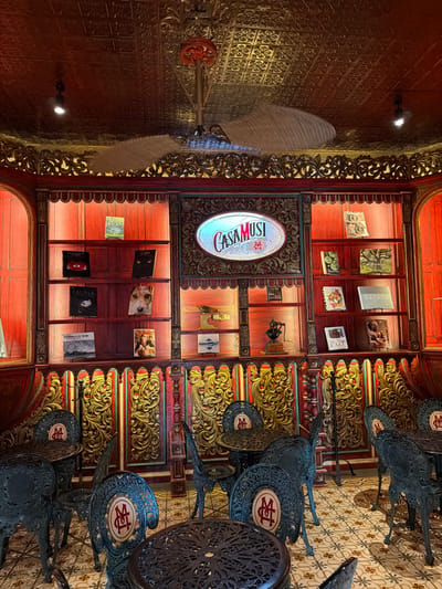 The interior features a cabinet display showcasing books and decorative items under warm lighting, framed by ornate wood detailing. Ceiling panels create a patterned backdrop. Vintage cast-iron tables and chairs sit on a tiled floor. A large fan hangs from the ceiling. The "Casa Musi" logo is prominently displayed within the cabinet.