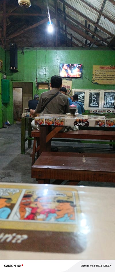 The image shows a scene from inside a small restaurant in Pasuruan, Indonesia, located at coordinates -7.663, 112.906. Three men are seated at a table, with one man at the far end wearing a brown shirt and a black strap across his shoulder. The man in the brown shirt appears to be the only one actively engaged in conversation. The other two men are looking down at the table or at their phones. The restaurant is simply furnished with wooden tables and benches. The walls are painted green and decorated with posters. There is a television screen on the wall playing what appears to be a boxing match. The restaurant is lit by a ceiling fan and a single light bulb. The table is covered with a brown tablecloth and there is a small glass with a red liquid on the table next to the man in the brown shirt. In the foreground, the image shows a close-up of a wooden table covered with a colorful tablecloth. The table has a cartoon-style illustration on it with the words "Sobat Warjo" printed in the center. The camera is slightly tilted, giving the impression that the person taking the picture is seated at the table in the foreground.