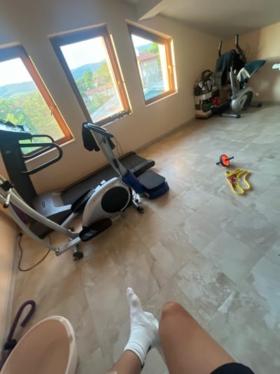The image shows an indoor home gym with exercise equipment, including a treadmill and an elliptical machine. The room has a tiled floor and three skylight windows that offer a view of a lush, green landscape with mountains in the distance, suggesting a daytime setting. Sunlight streams through the windows, illuminating the space and casting shadows.

On the floor, a bright yellow resistance band and an orange abdominal roller are visible. In the background, a shelf unit is stacked with various items, including what appear to be books and storage containers. A person's legs, wearing white socks and dark athletic shorts, are partially visible in the foreground, indicating they are likely using the gym. The overall impression is a functional and somewhat cluttered home fitness area bathed in natural light. The location cues from the windows point to Krainitsi, Bulgaria. There is no visible text in the image.