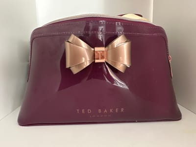 This is a close-up, eye-level, frontal shot of a shiny, deep red cosmetic bag. The bag is rectangular with rounded edges and a zippered closure at the top, which has a silver-tone zipper. Adorning the front of the bag is a large, satin-finish, rose gold bow with a rose gold "T" emblem in the center. Below the bow, the brand name "TED BAKER LONDON" is printed in rose gold lettering. The bag sits on a white surface, possibly a shelf, with a white wall visible in the background. The lighting suggests an indoor setting, likely during the daytime. There is no activity taking place; the image is a still life showcasing the product. The focus is entirely on the cosmetic bag, highlighting its material, design, and branding.